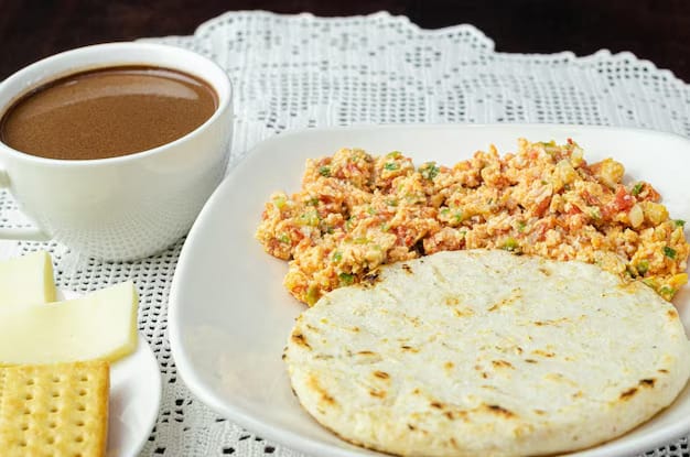 A typical Colombian breakfast plate featuring a white corn arepa, fluffy huevos pericos (scrambled eggs with tomato), and a cup of coffee.
