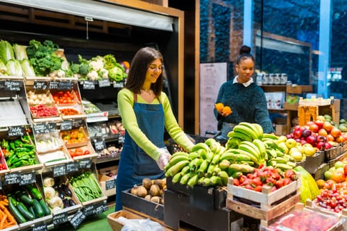 A woman selecting fresh green plantains at a Colombian market, illustrating that the foundation of the cuisine answers the question 'is Colombian food spicy?' with a focus on fresh ingredients, not peppers.