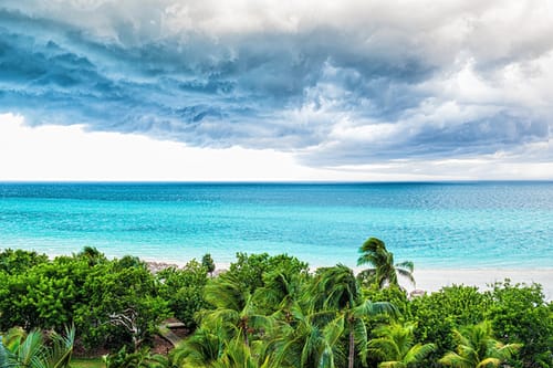 Dramatic storm clouds gathering over the vibrant turquoise Caribbean Sea, with the lush palm trees of the Colombian islands bending in the wind.