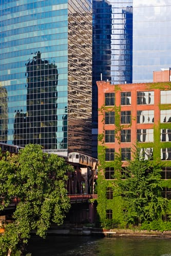 A juxtaposition of modern glass skyscrapers reflecting the urban environment and an older brick building covered in ivy, with trees in the foreground. This image highlights the architectural changes and development often seen during gentrification in Medellin.