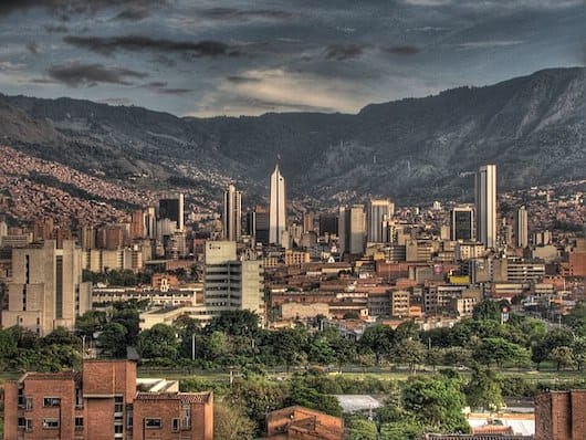 An expansive cityscape of Medellin with numerous high-rise buildings and scattered green areas, framed by dramatic, cloud-covered mountains in the background, showcasing the contrast between urban life and the natural beauty available on nature day trips from Medellin.
